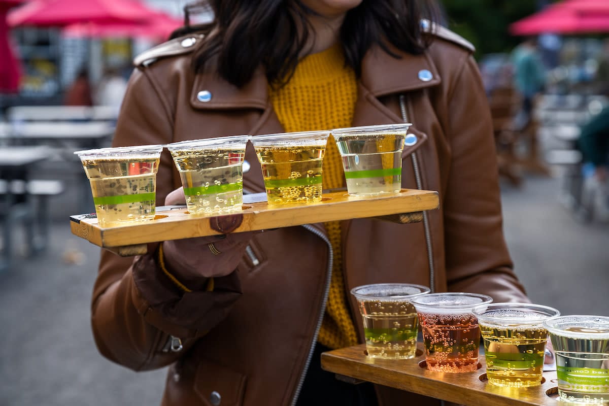 At the NYC Cider Festival, a person in a brown jacket holds a tray laden with plastic cups brimming with an array of beers and ciders. The drinks, ranging from light to amber hues, complement the lively outdoor seating adorned with red umbrellas in the background.