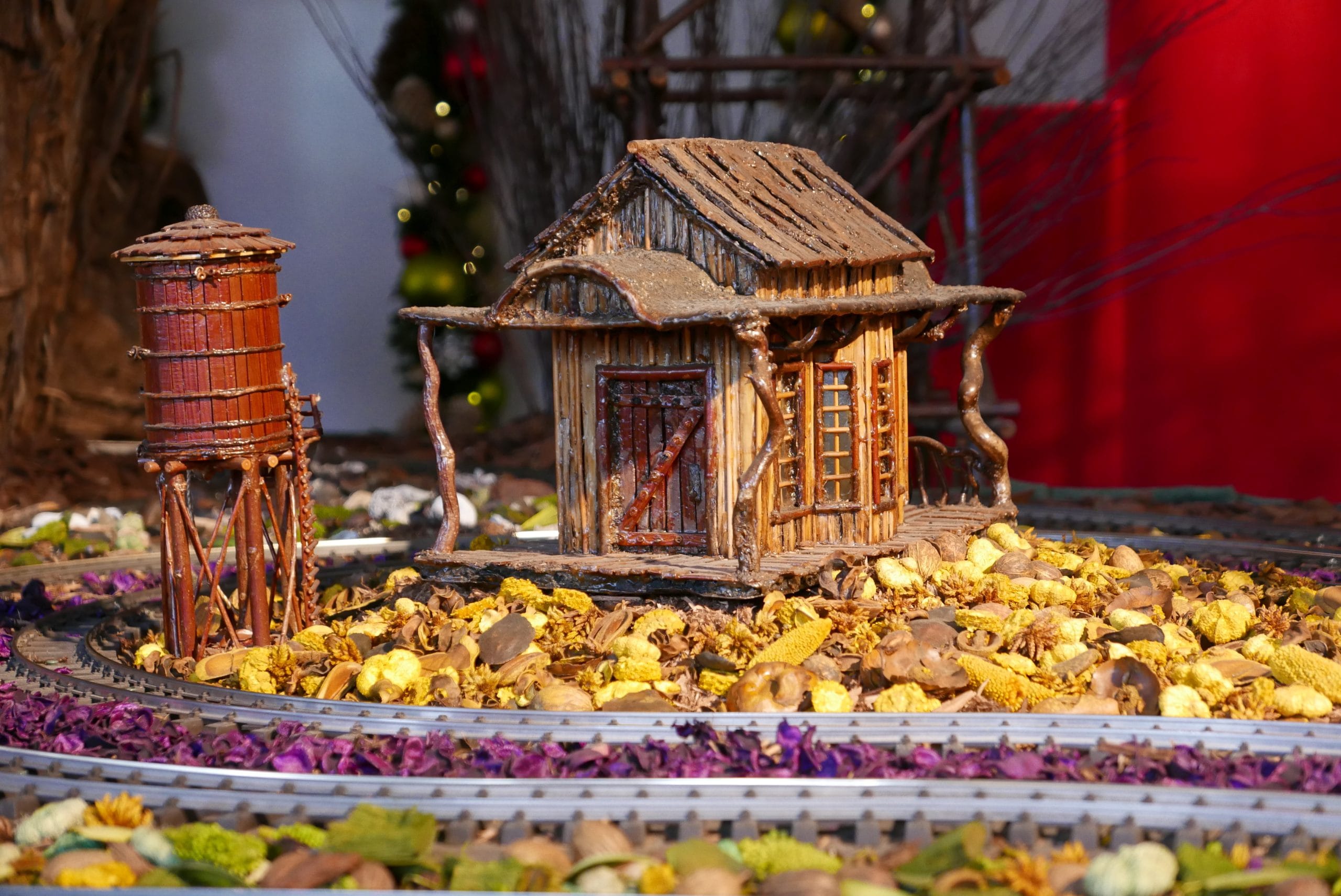 A small model house and wooden water tower sit on a circular track surrounded by dried leaves, flowers, and stones, evoking the charm of the New York Botanical Garden Holiday Train Show. The blurred background features festive decorations. A small model house and wooden water tower sit on a circular track surrounded by dried leaves, flowers, and stones, evoking the charm of the New York Botanical Garden Holiday Train Show. The blurred background features festive decorations.