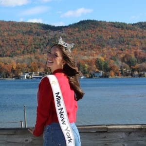 Abigail Quammen seen wearing a red sweater and Miss New York sash smiles by Lake George, crowned with elegance. The background features autumn-colored trees and a hillside under the blue sky, capturing the serene beauty of this iconic location.