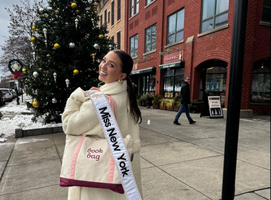 Abigail Quammen, Miss New York, stands on a snowy Saratoga sidewalk, smiling and holding a tote bag labeled book bag. She is proudly wearing her Miss New York sash draped over her shoulder. A decorated Christmas tree and charming brick buildings complete the festive backdrop.