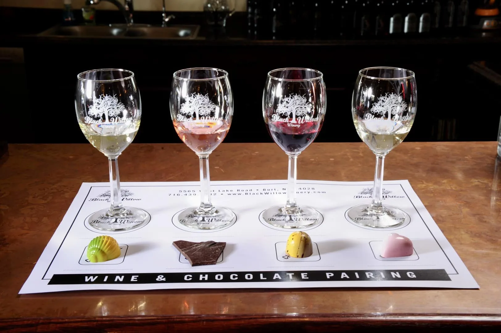 Four wine glasses filled with different types of wine are arranged on a menu labeled Wine & Chocolate Pairing at the Capital Wine Festival. In front of each glass is a unique chocolate piece, all elegantly set on a bar with a dark wood counter.