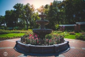 A tiered stone fountain stands surrounded by blooming pink flowers and a black iron fence in a sunny, landscaped Schenectady park with trees and brick pathways.