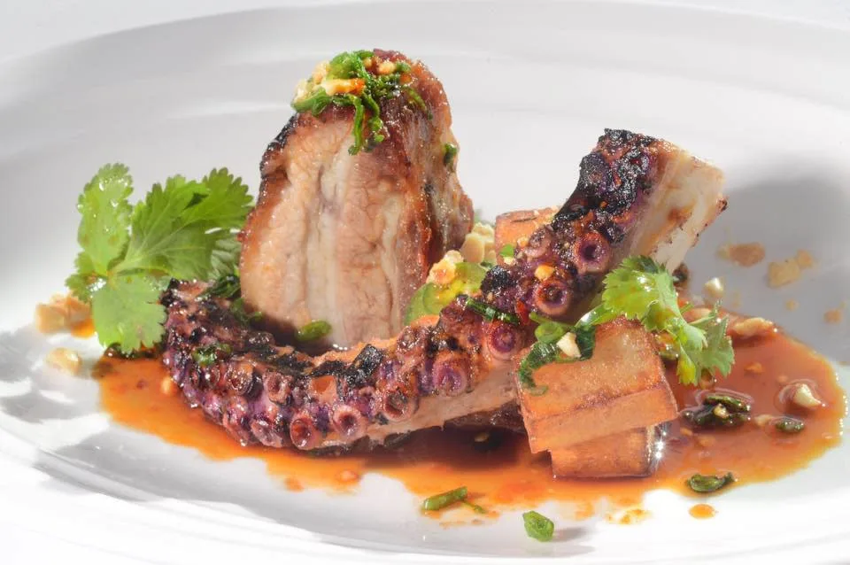 Grilled octopus tentacle and pork belly, served with sauce, fresh cilantro, diced herbs, and golden roasted potato cubes on a white plate—a standout dish in the heart of Saratoga Springs vibrant Restaurant City at 15 Church