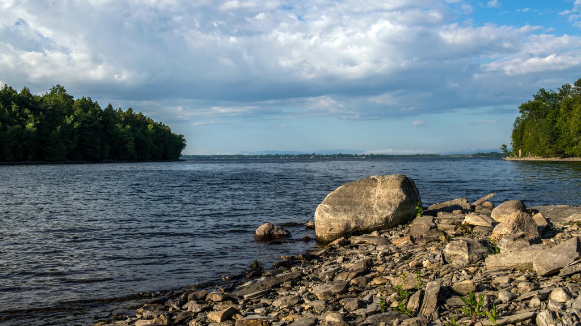 Rocky shoreline with a large boulder in the foreground along Plattsburghs calm river, green trees lining both sides. The sky above is partly cloudy with patches of blue.