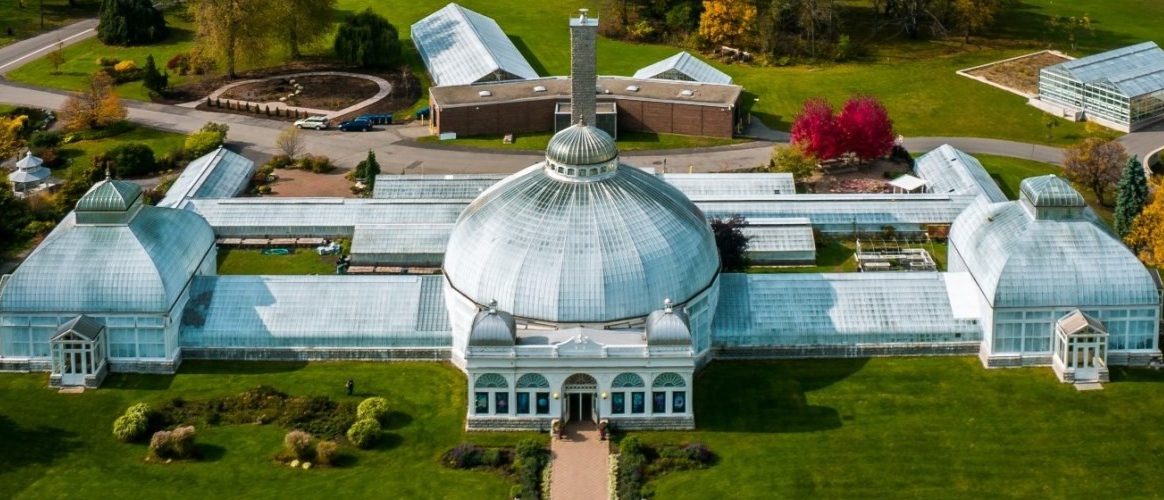 Aerial view of a large glass conservatory with a central dome and attached greenhouses in Buffalo, surrounded by green lawns, trees, and nearby buildings.