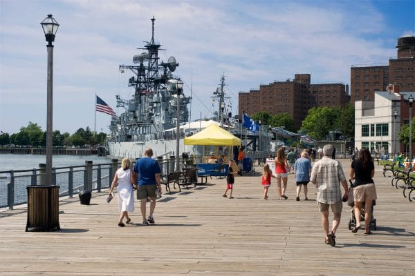 People stroll along a wooden boardwalk beside the river in Buffalo, with a large naval ship docked nearby. Buildings, benches, and a yellow tent dot the sunny scene, while an American flag waves proudly from the ship.