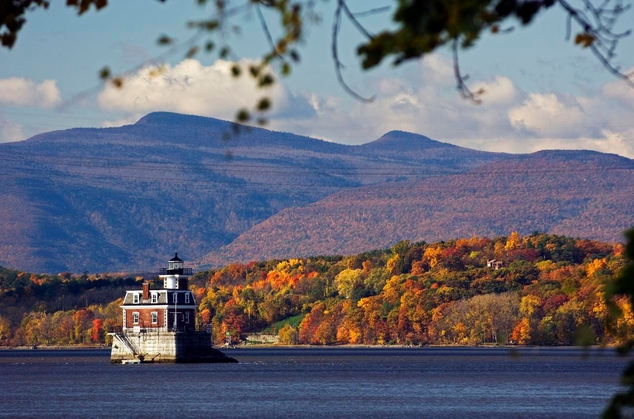 A small lighthouse stands on a stone base in a wide river, surrounded by autumn trees in vivid shades of orange, red, and yellow—an idyllic scene perfect for car-free travel or scenic trips by train through blue mountains under a partly cloudy sky.