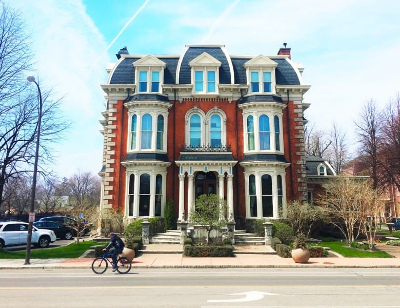 A large, ornate red brick Victorian mansion in Buffalo with tall windows, decorative trim, and two potted shrubs at the entrance. A person rides a bicycle on the street in front, and cars are parked along the sidewalk.