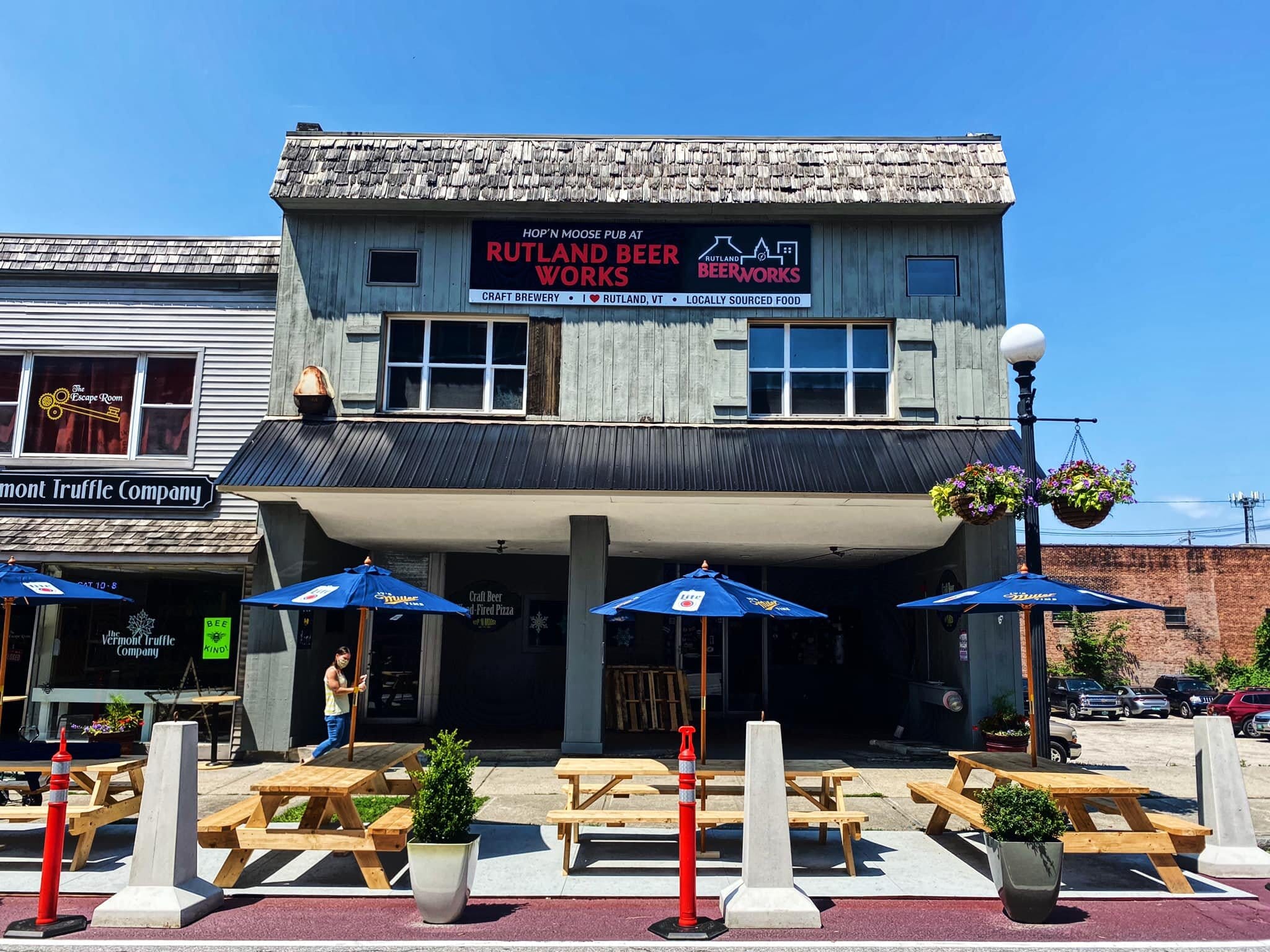 Street view of Rutland Beer Works pub in downtown Rutland, featuring covered outdoor seating with blue umbrellas, picnic tables, and hanging flower baskets; nearby shops include a chocolate store. Clear, sunny day.