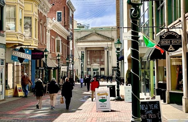 A lively pedestrian street in Schenectady with people strolling past shops, including an Irish shop with a hanging sign and flag. Historic brick buildings line the way, leading to a grand, columned structure in the background.