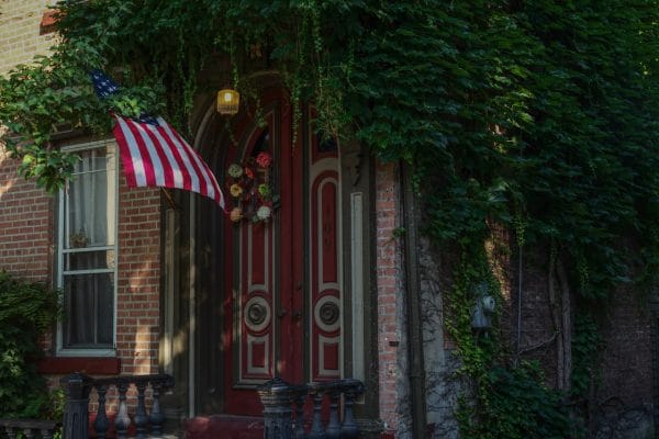A red double door with floral wreaths and an arched window is surrounded by lush green vines in Schenectady. An American flag hangs beside the door, with a small lantern glowing above, set against brick walls and a visible window.