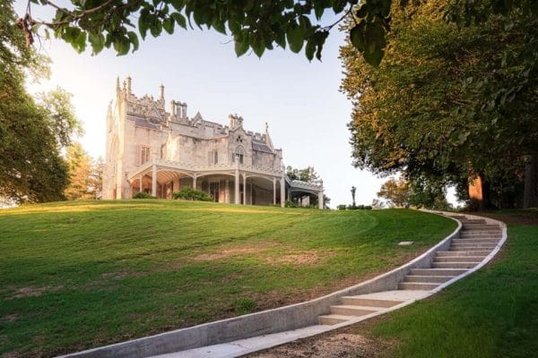 A historic mansion with ornate architecture, reminiscent of the legendary Crafts at Lyndhurst, sits atop a grassy hill surrounded by large trees. A curved staircase leads up the hill, bordered by lush greenery under a clear sky.