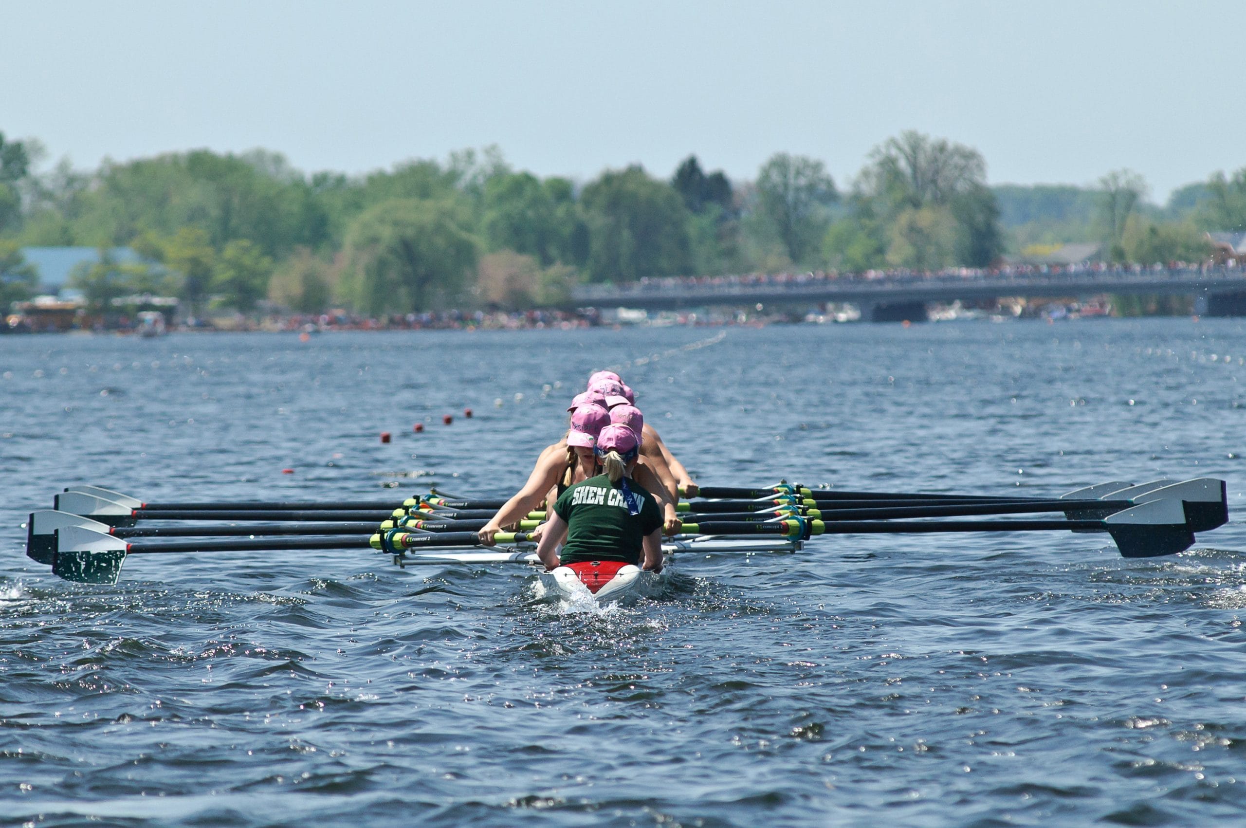 A rowing team, sporting matching uniforms, paddles in unison at the Saratoga Invitational Rowing Regatta. They glide across a calm body of water towards a distant shoreline adorned with trees and a bridge, all under a pristine sky.