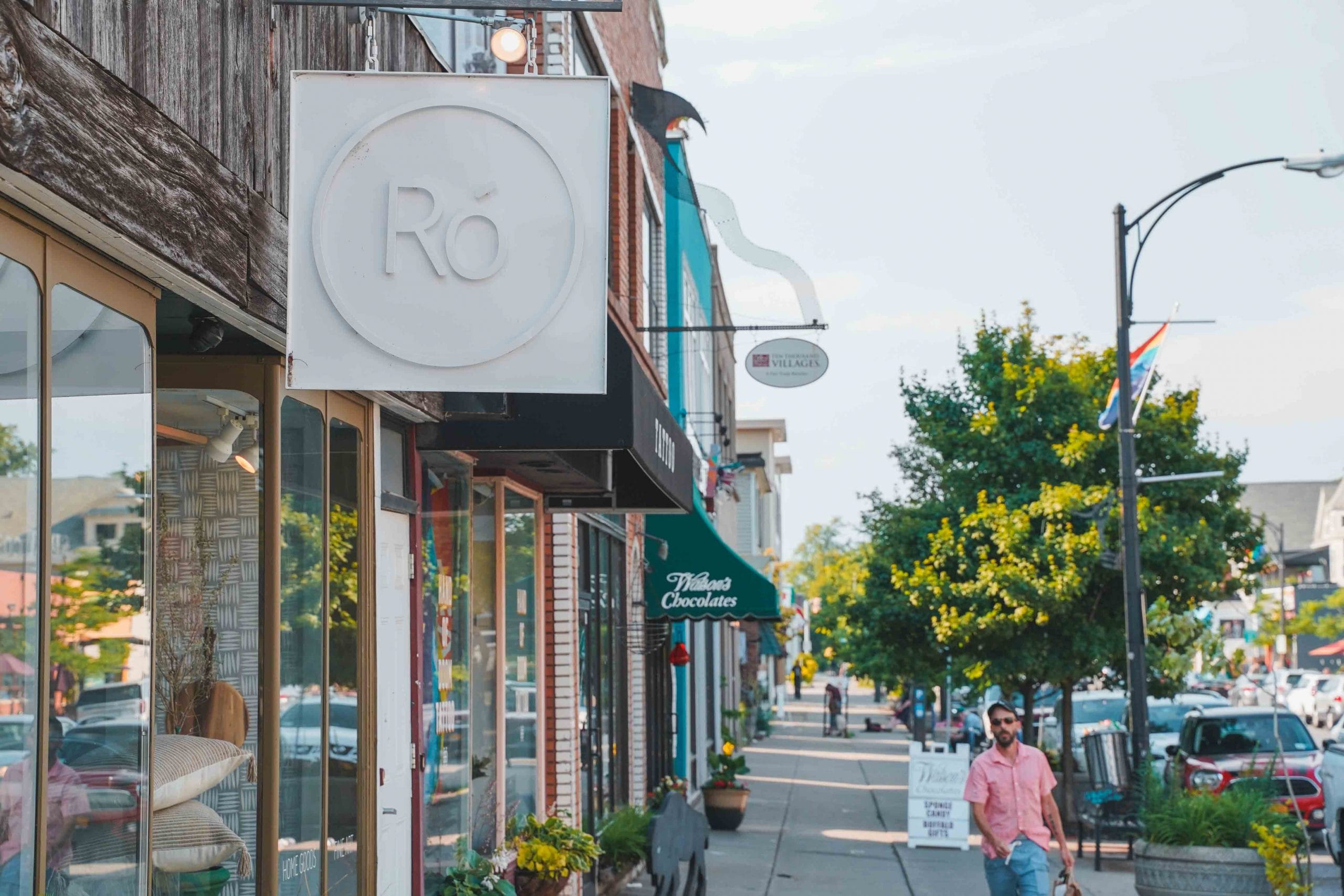 A man in a pink shirt walks down a lively street in Buffalo, lined with shops—including one with a sign reading Ró. Cars are parked along the sidewalk, and trees provide shade on a sunny day.