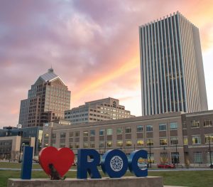 Large I ♥ ROC sculpture in front of modern Rochester office buildings at sunset, with dramatic pink and orange clouds overhead. The scene is set in a city plaza with grass and concrete walkways.