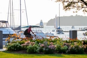 A person rides a bicycle past colorful flowers in the foreground, embracing car-free travel as boats and sailboats drift on a sunlit lake and a lighthouse rises in the background. Burlington Vermont Waterfront