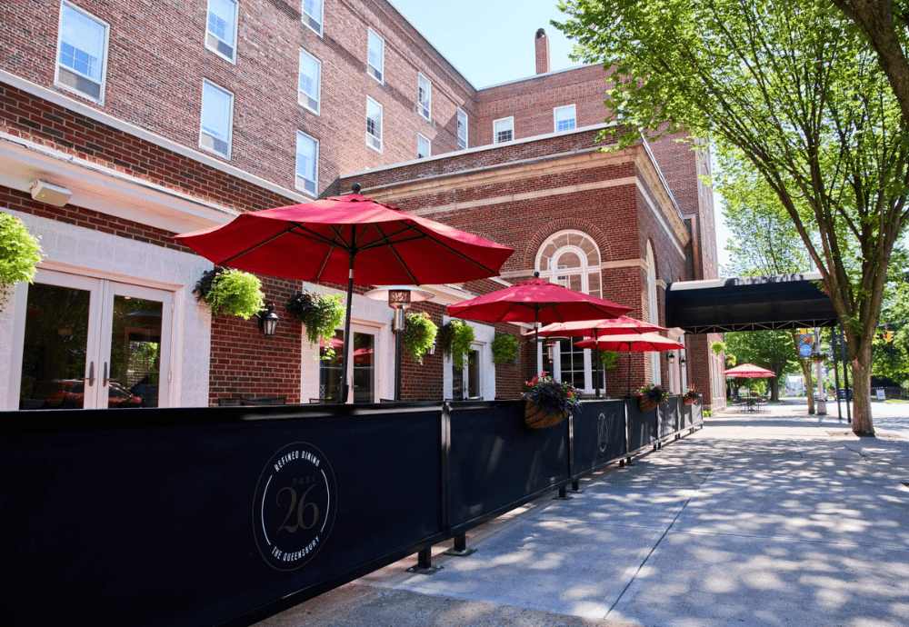 Outdoor restaurant patio beside a red-brick building, featuring red umbrellas over tables, hanging plants, and black railings with 26 logo; just steps from the Queensbury Hotel. Trees line the sidewalk under a sunny, blue sky.