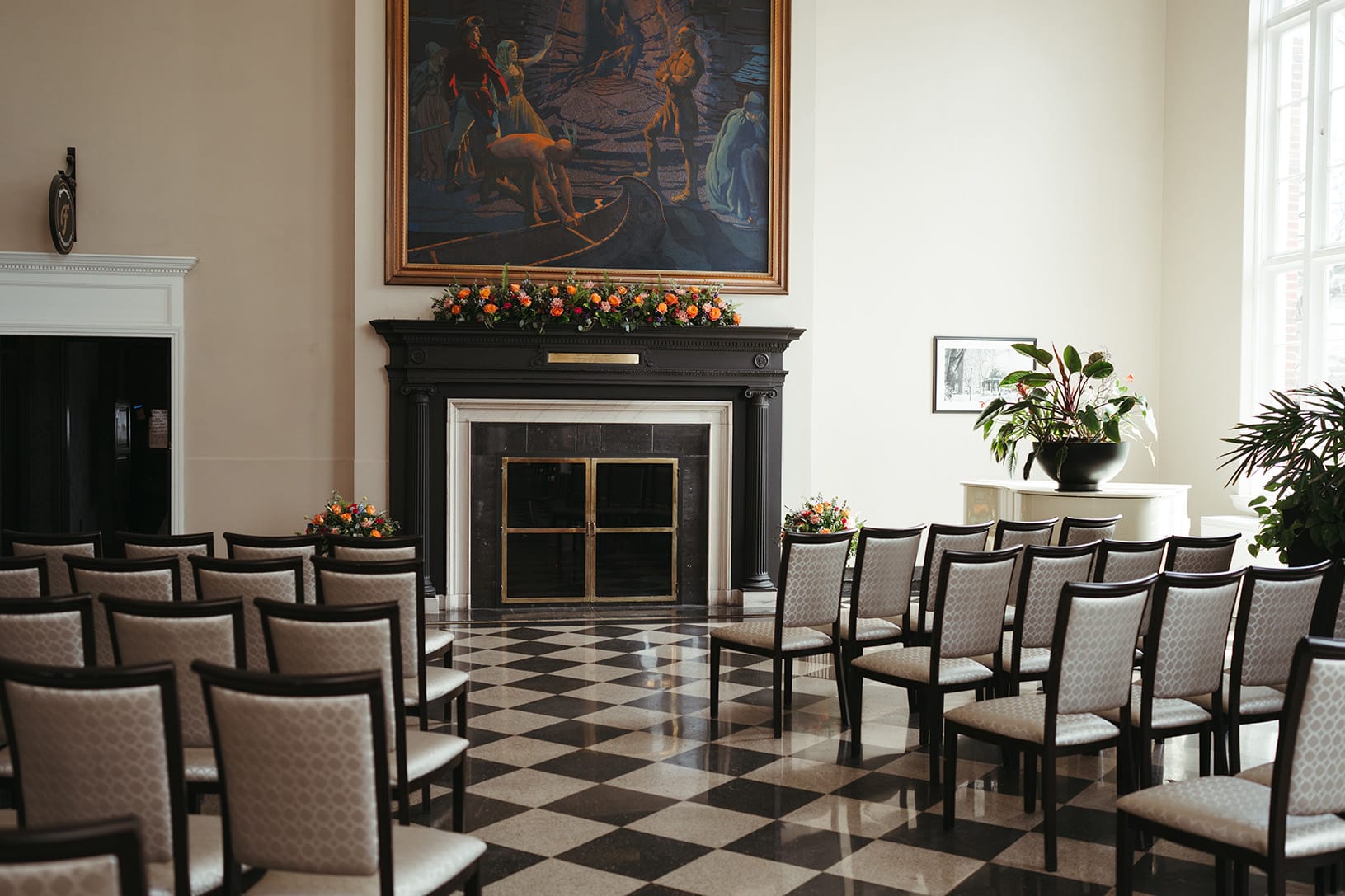 A room at the Queensbury Hotel with rows of empty chairs facing a flower-adorned fireplace. Above it hangs a large painting, while sunlight streams through tall windows onto the checkered black and white floor.
