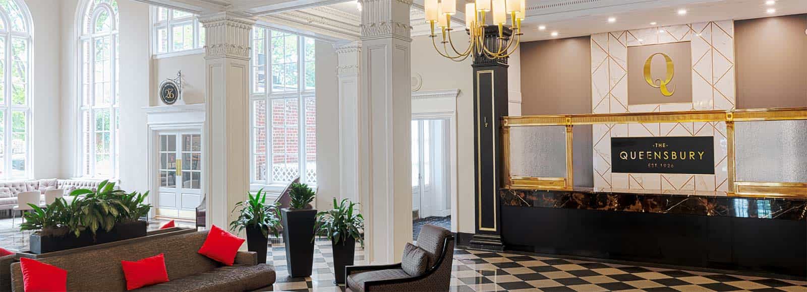 Bright Queensbury Hotel lobby with tall windows, white walls, black and white tiled floor, modern reception desk with “The Queensbury” sign, green plants, and a seating area featuring brown sofas and red cushions.