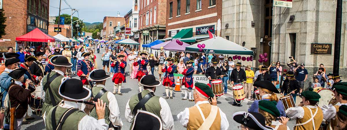 A lively Ticonderoga StreetFest parade features people in colonial-era costumes playing drums and flutes, surrounded by onlookers, tents, and historic brick buildings on a sunny day.