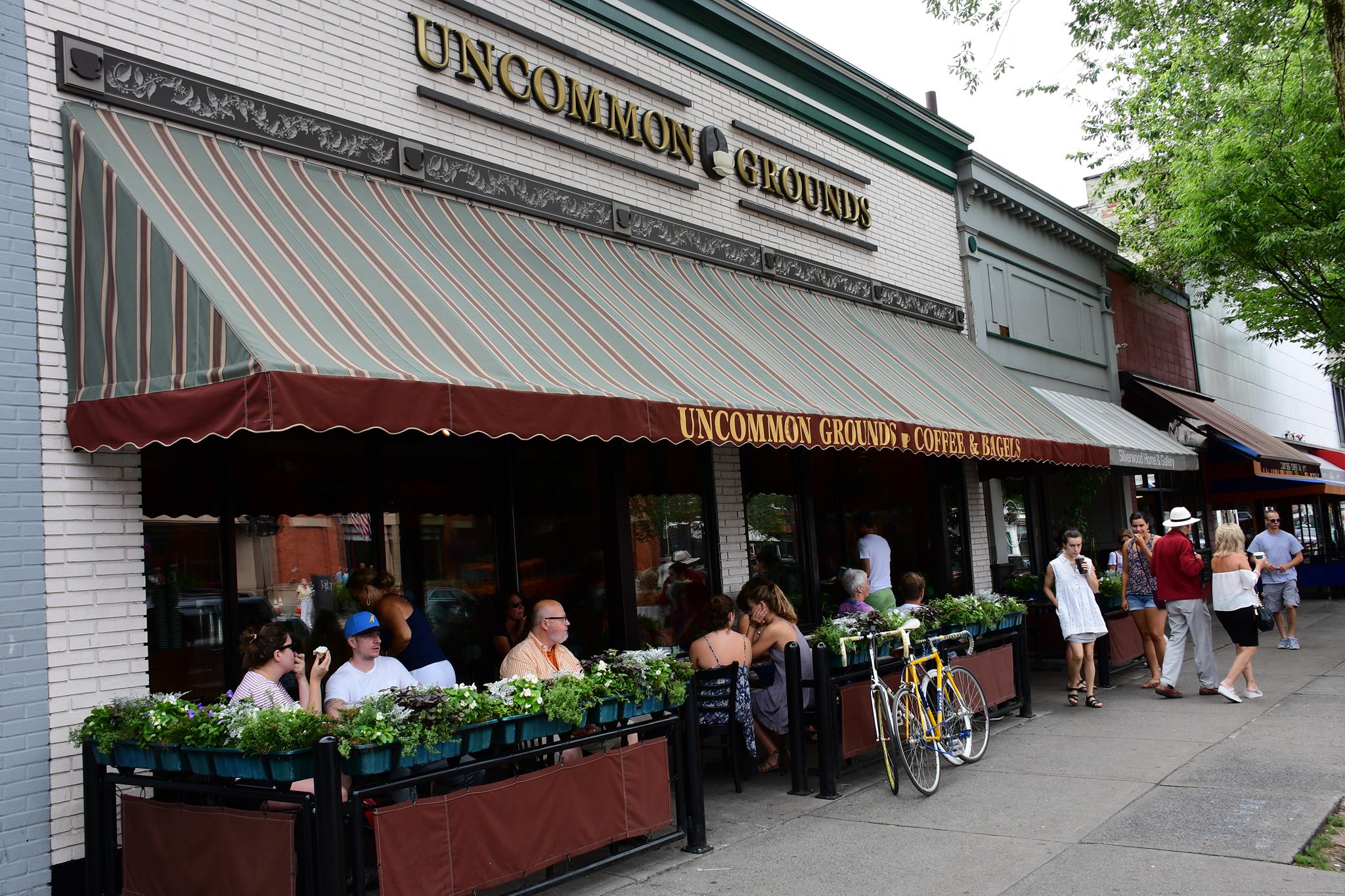People sit at outdoor tables under a striped awning at Uncommon Grounds. Flower boxes line the patio, a yellow bicycle is parked nearby, and pedestrians stroll along the sidewalk in front of the cozy café.