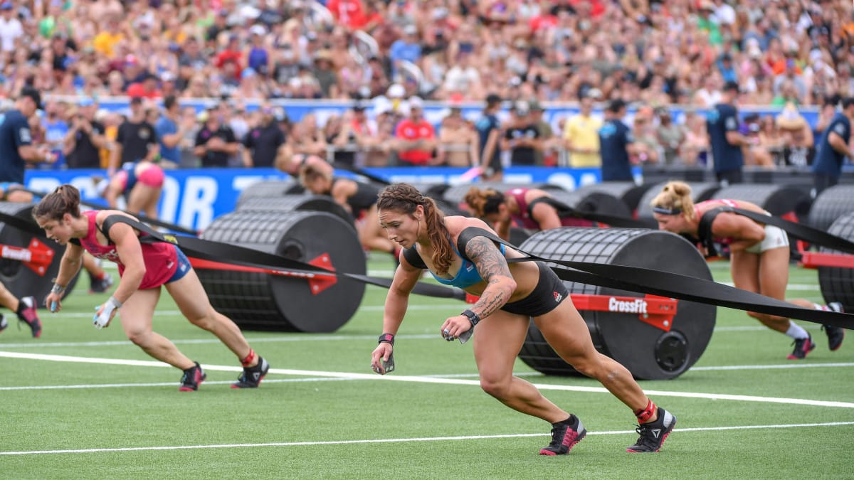 Several women compete in a CrossFit Games-style strongman event, pulling large weighted sleds across a field while a crowd watches in the background. The athletes appear focused and determined.