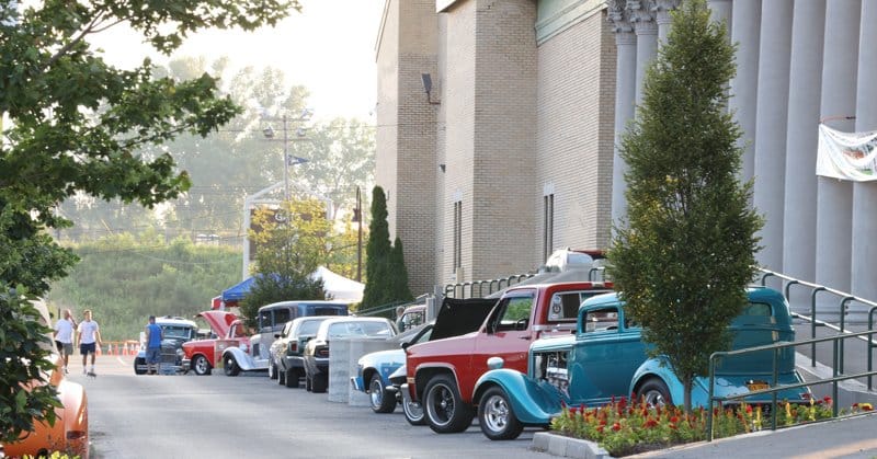 Classic cars and vintage trucks are parked in a row along the side of a building with columns during the Blues Festival; people walk in the background, and trees line the street on a sunny day.