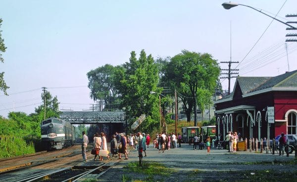 A vintage passenger train with historic roots approaches a busy small-town station where people with suitcases are waiting or greeting arrivals near a red building and trees on a sunny day.