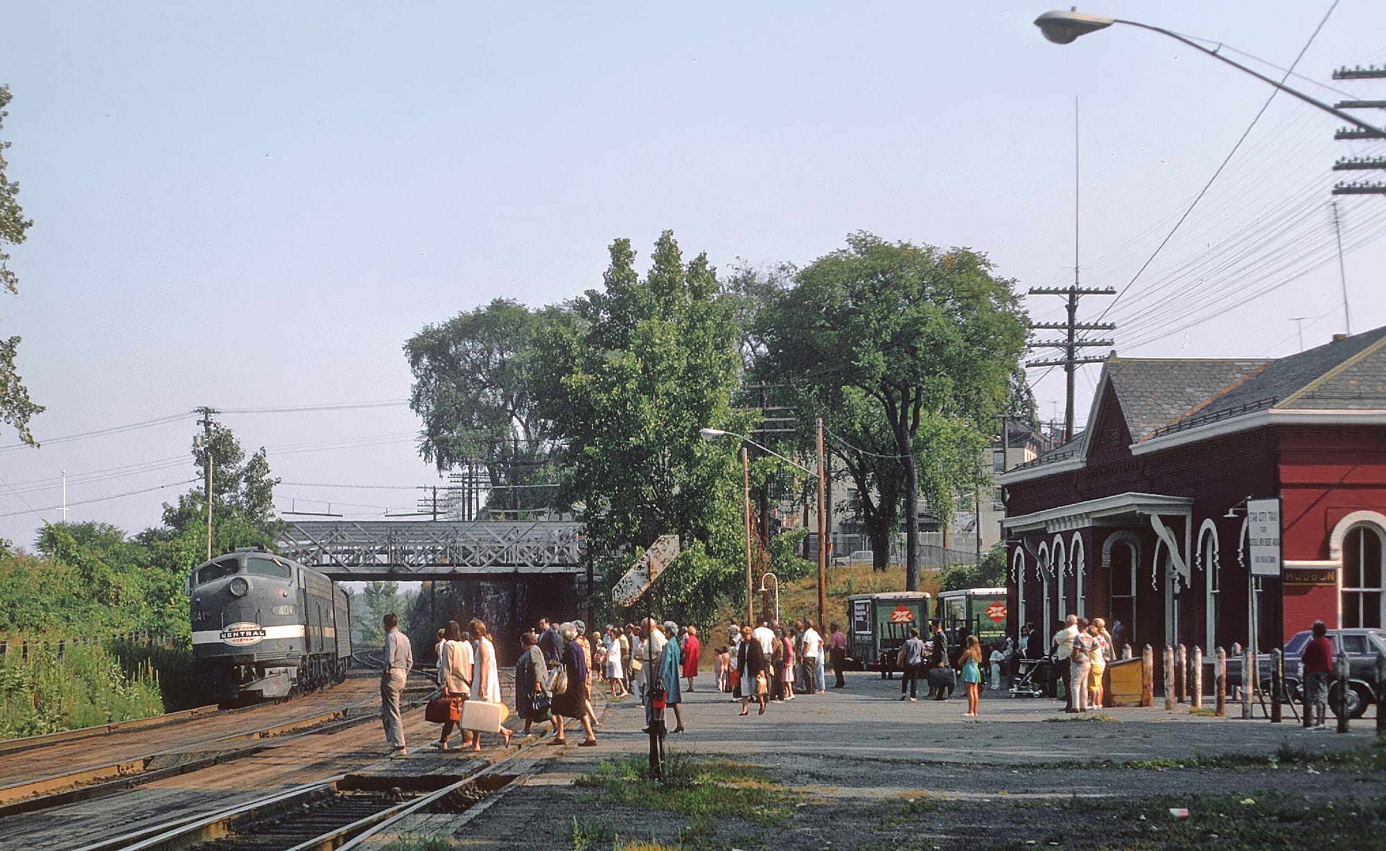A vintage passenger train with historic roots approaches a busy small-town station where people with suitcases are waiting or greeting arrivals near a red building and trees on a sunny day.