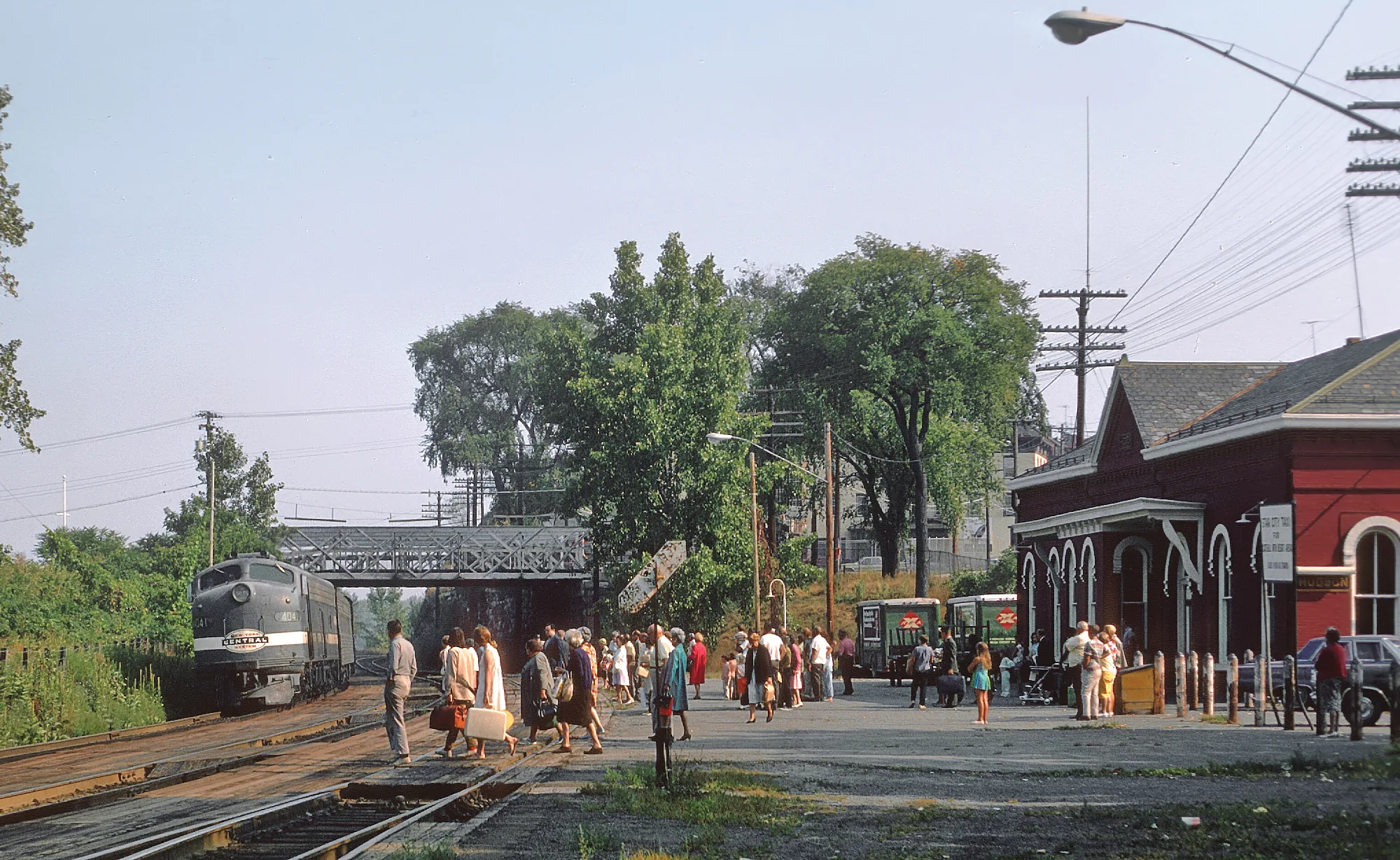 A vintage passenger train with historic roots approaches a busy small-town station where people with suitcases are waiting or greeting arrivals near a red building and trees on a sunny day.