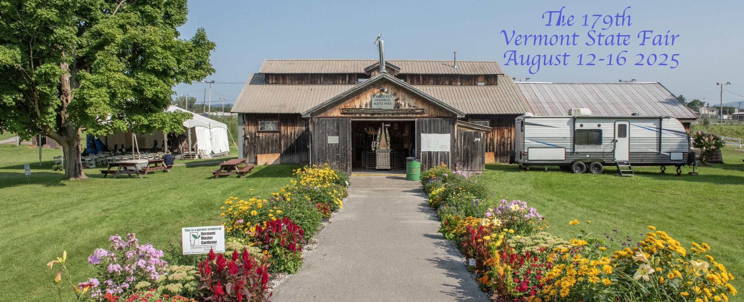 A wooden building at the Vermont State Fair, with colorful flowers bordering the walkway, a tree and tents on the left, and an RV on the right. Text reads: “The 179th Vermont State Fair August 12–16, 2025.”.