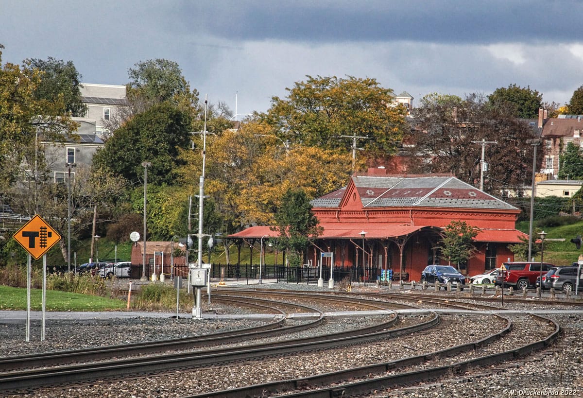 A curved railroad track leads to a historic red train station with deep historic roots, surrounded by trees with autumn foliage. Several cars are parked nearby and a yellow railroad crossing sign is visible in the foreground.