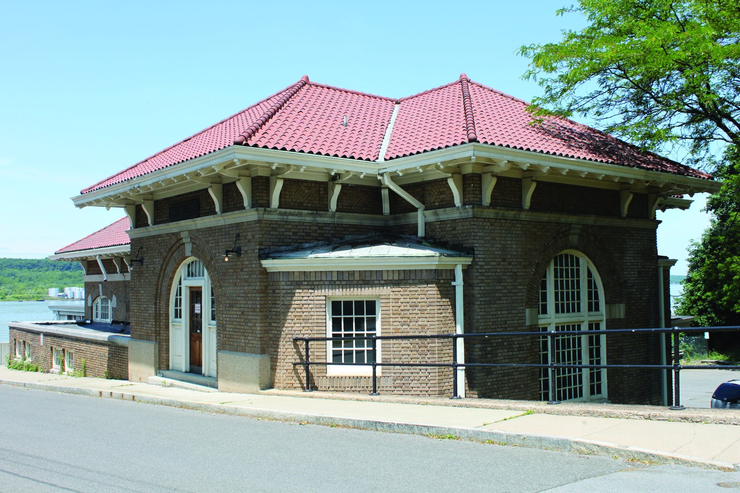 Historic brick building with a red-tiled roof and arched windows, situated on a slightly sloped street with a handrail and sidewalk in front. Its historic roots shine through, with trees and a river visible in the background.