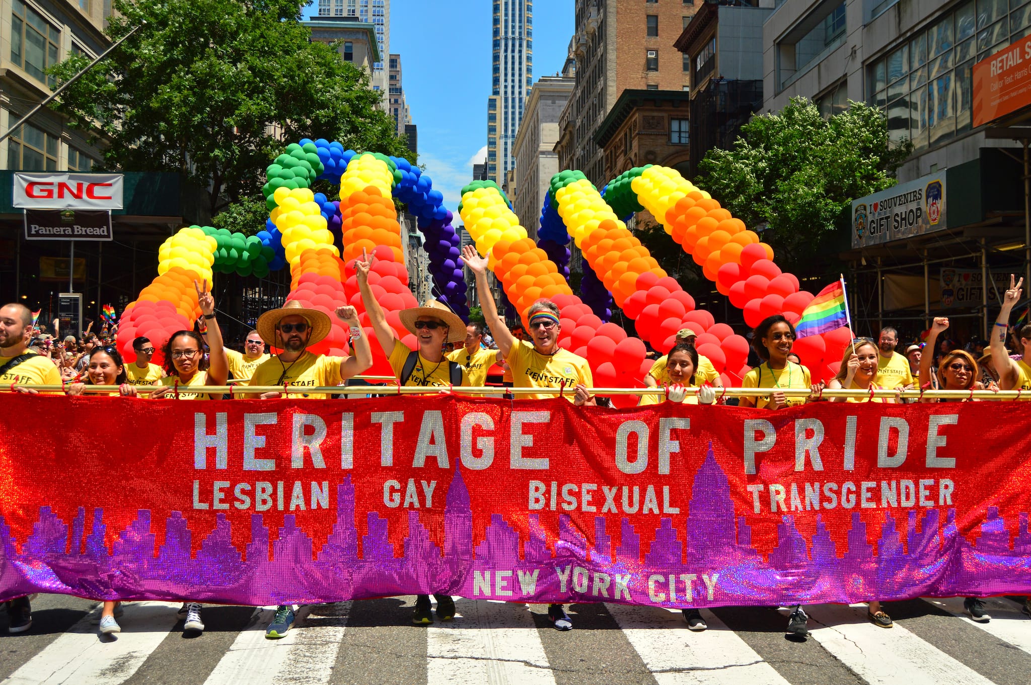 People march in a New York City Pride Parade holding a large red and purple banner reading “Heritage of Pride: Lesbian, Gay, Bisexual, Transgender.” Colorful rainbow balloons arch above the proud group as they walk down a city street.