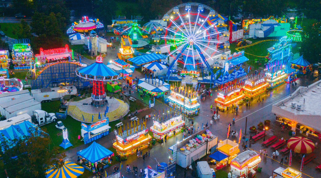 A vibrant aerial view of the Dutchess County Fair at night, featuring a large Ferris wheel, bright lights, various rides, food stands, and people walking along wet pathways.