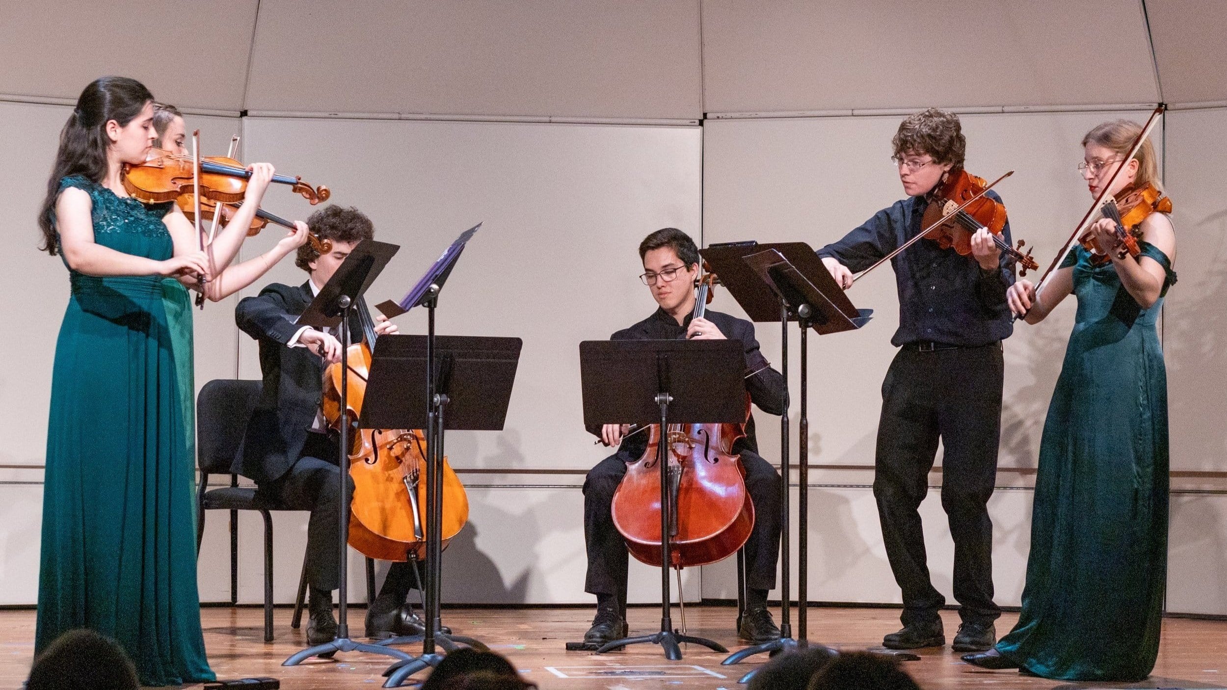 Five musicians perform on stage at the Manchester Music Festival; three women in matching green dresses play violins and viola, while two men in black attire play cello and violin, all focused on their sheet music in a concert setting.