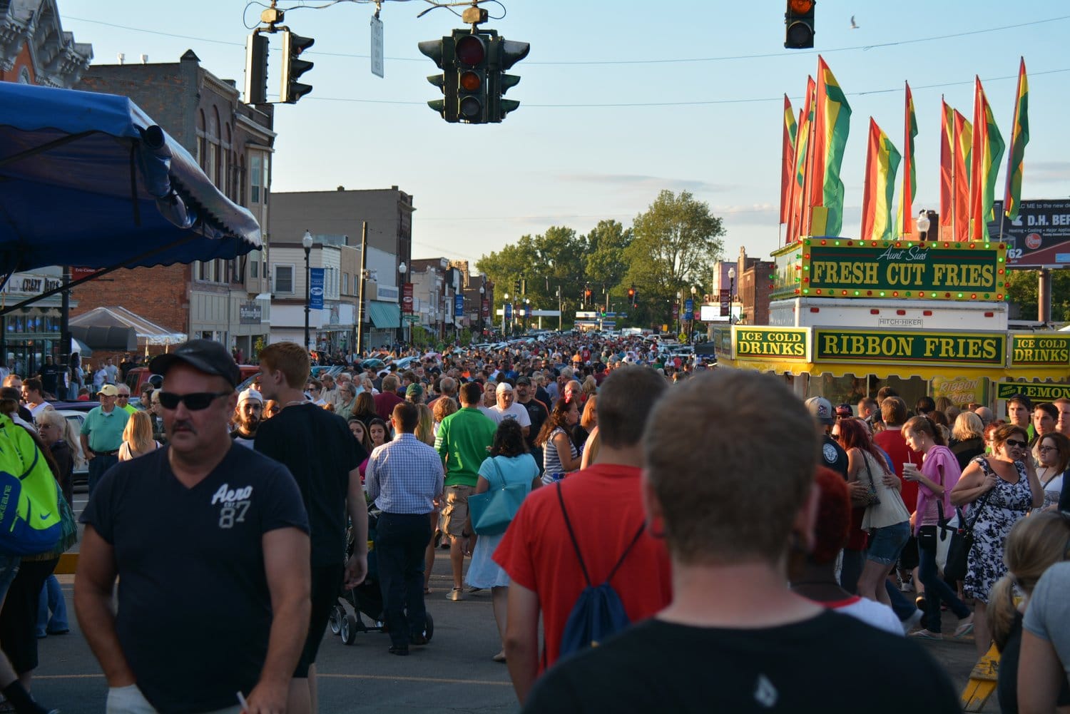 A large crowd of people walking through Canal Fest, a busy street fair with colorful flags, food stands offering fresh-cut ribbon fries and cold drinks; buildings and traffic lights line the street under a clear sky.