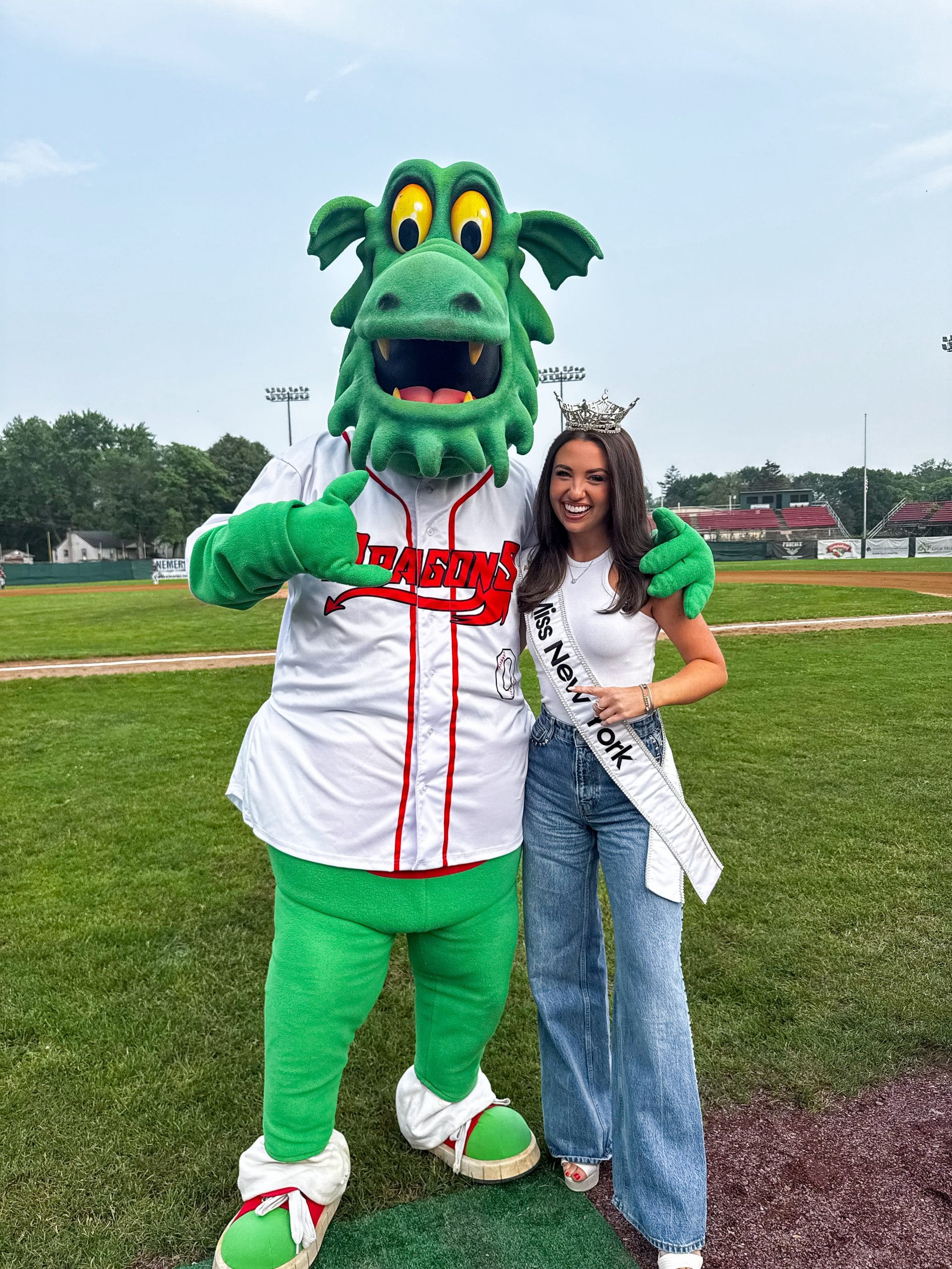 A woman wearing a crown and Miss New York sash stands on a baseball field in Lake George, smiling and posing with a person in a green dragon mascot costume in a Dragons jersey. Trees and stadium lights are visible in the background.