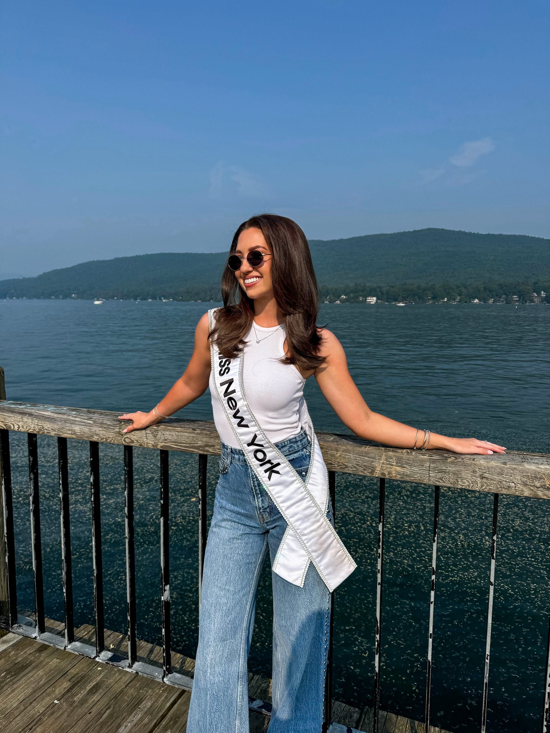 A woman wearing a Miss New York sash, sunglasses, a white top, and blue jeans leans on a wooden railing by the calm waters of Lake George, with green hills and a blue sky in the background.