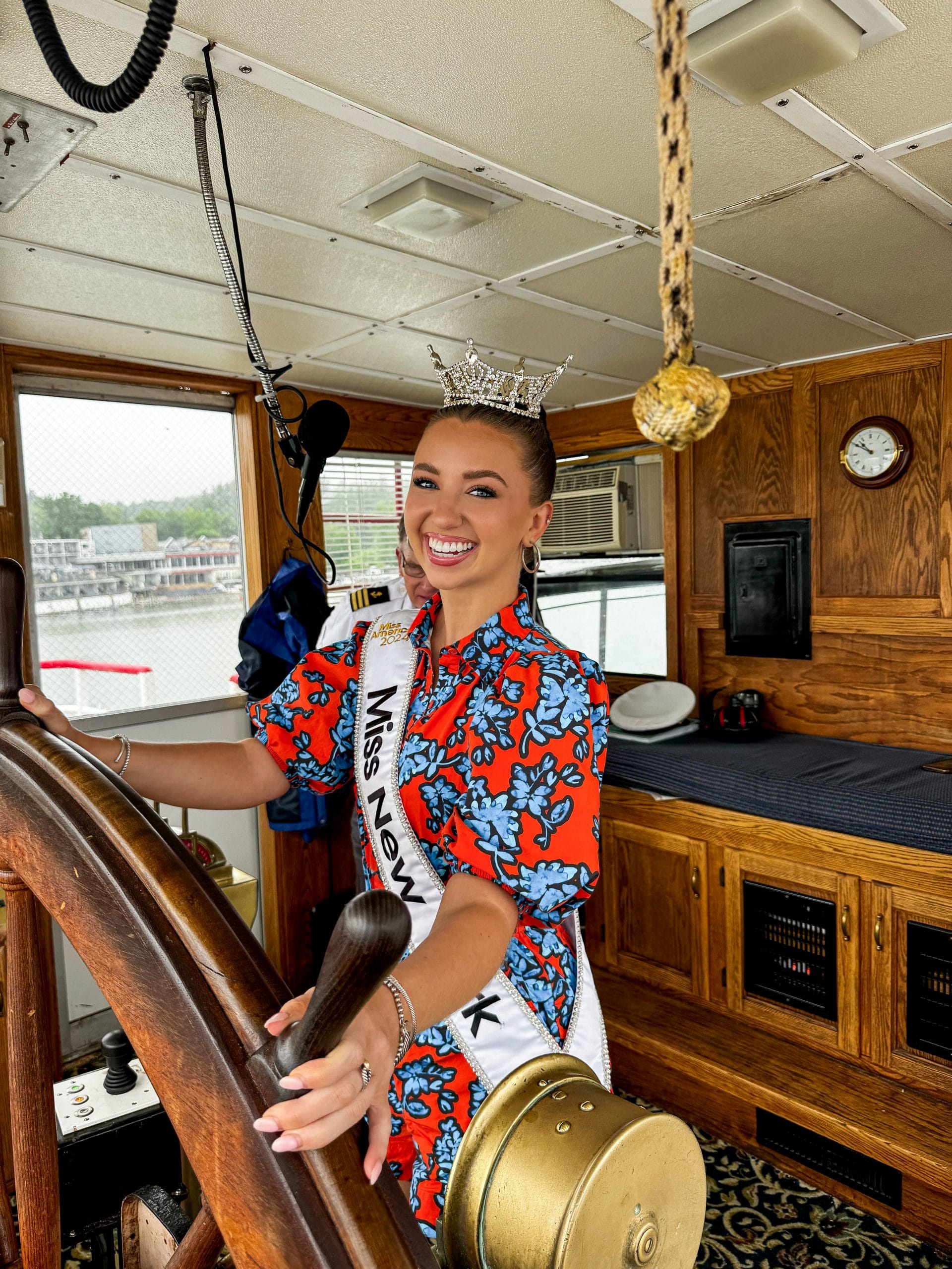 A woman wearing a crown and a Miss New York sash smiles while steering a wooden ship’s wheel inside a boat’s cabin on Lake George, dressed in a colorful floral dress.