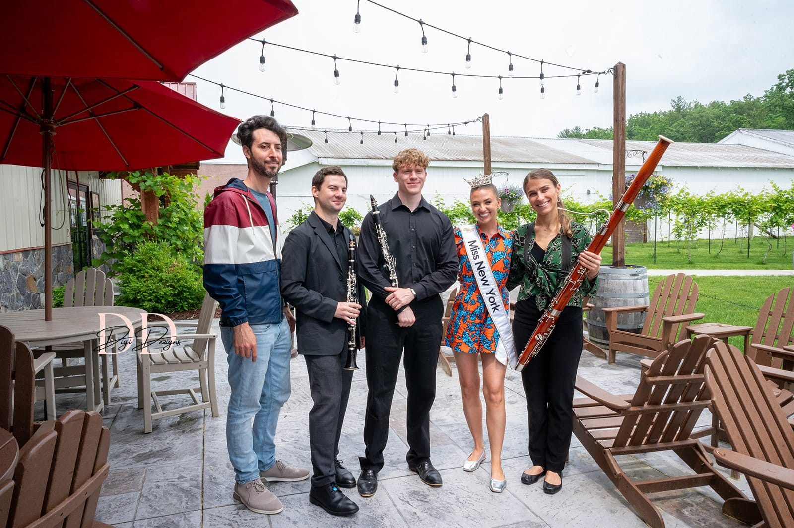 Five people stand outdoors on a patio near Lake George; three hold musical instruments while one wears a Miss New York sash. String lights hang overhead, surrounded by wooden chairs, a red umbrella, and lush greenery in the background.