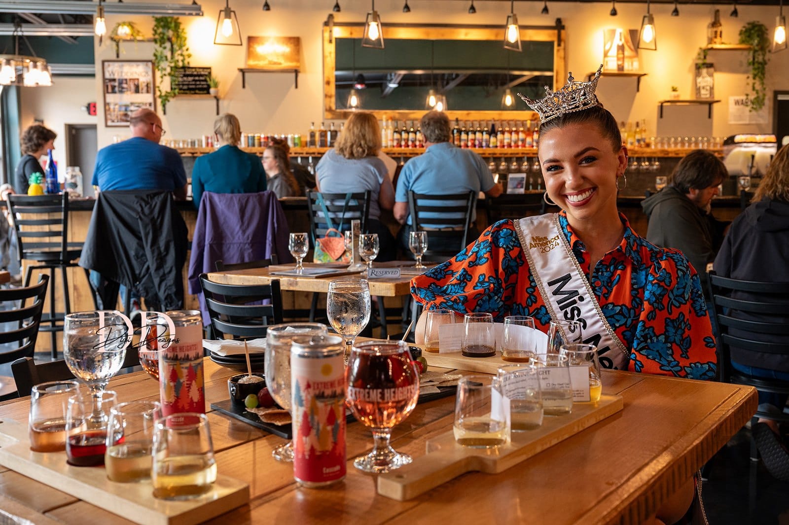 A woman wearing a Miss sash and crown smiles at a table with flight trays, glasses, and cans in a lively Lake George bar or brewery, with patrons seated at the bar in the background.