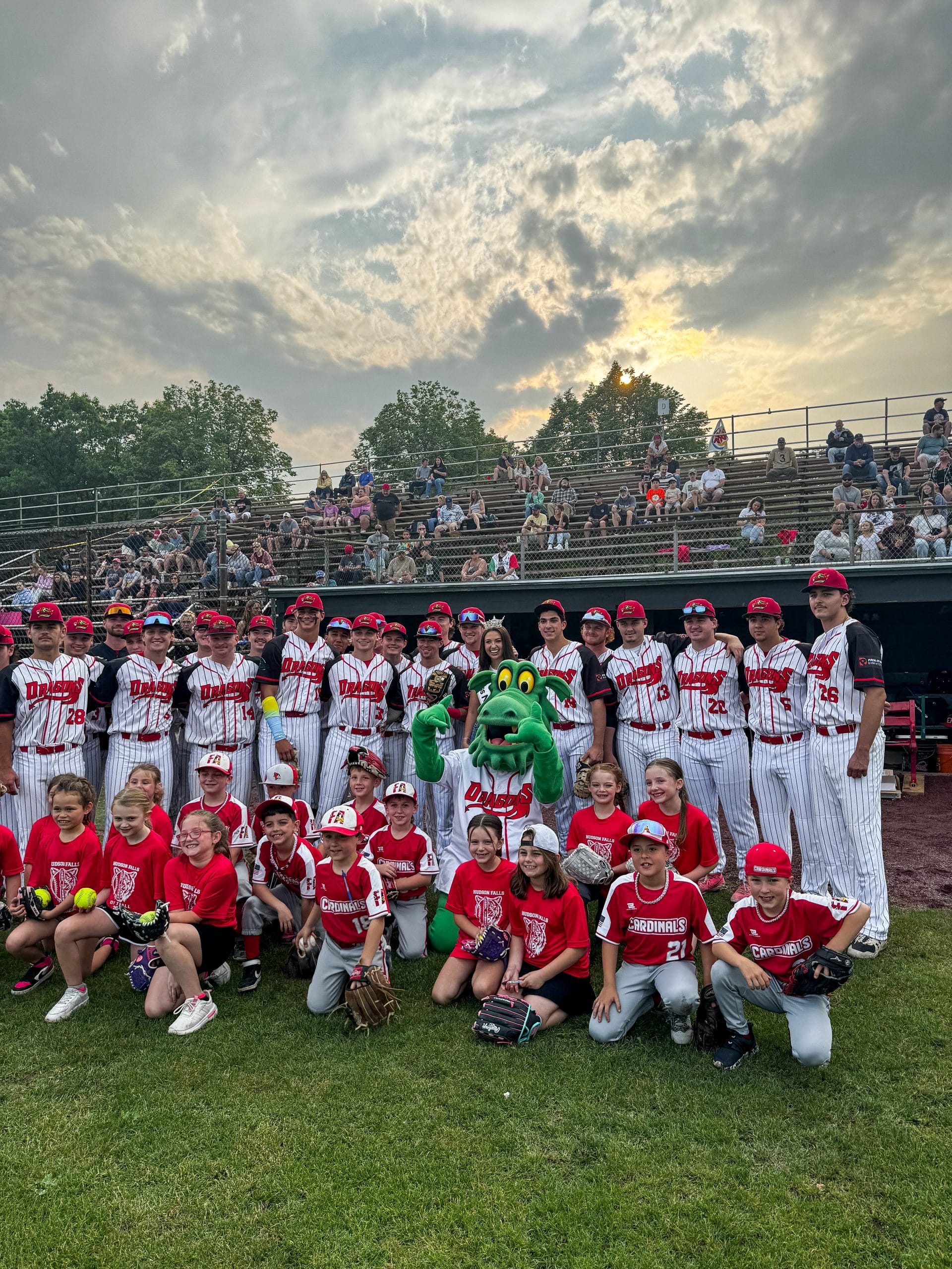A youth baseball team poses on a Lake George field with coaches, a green dragon mascot, and children in matching uniforms. Spectators fill the bleachers behind them under a cloudy sunset sky.