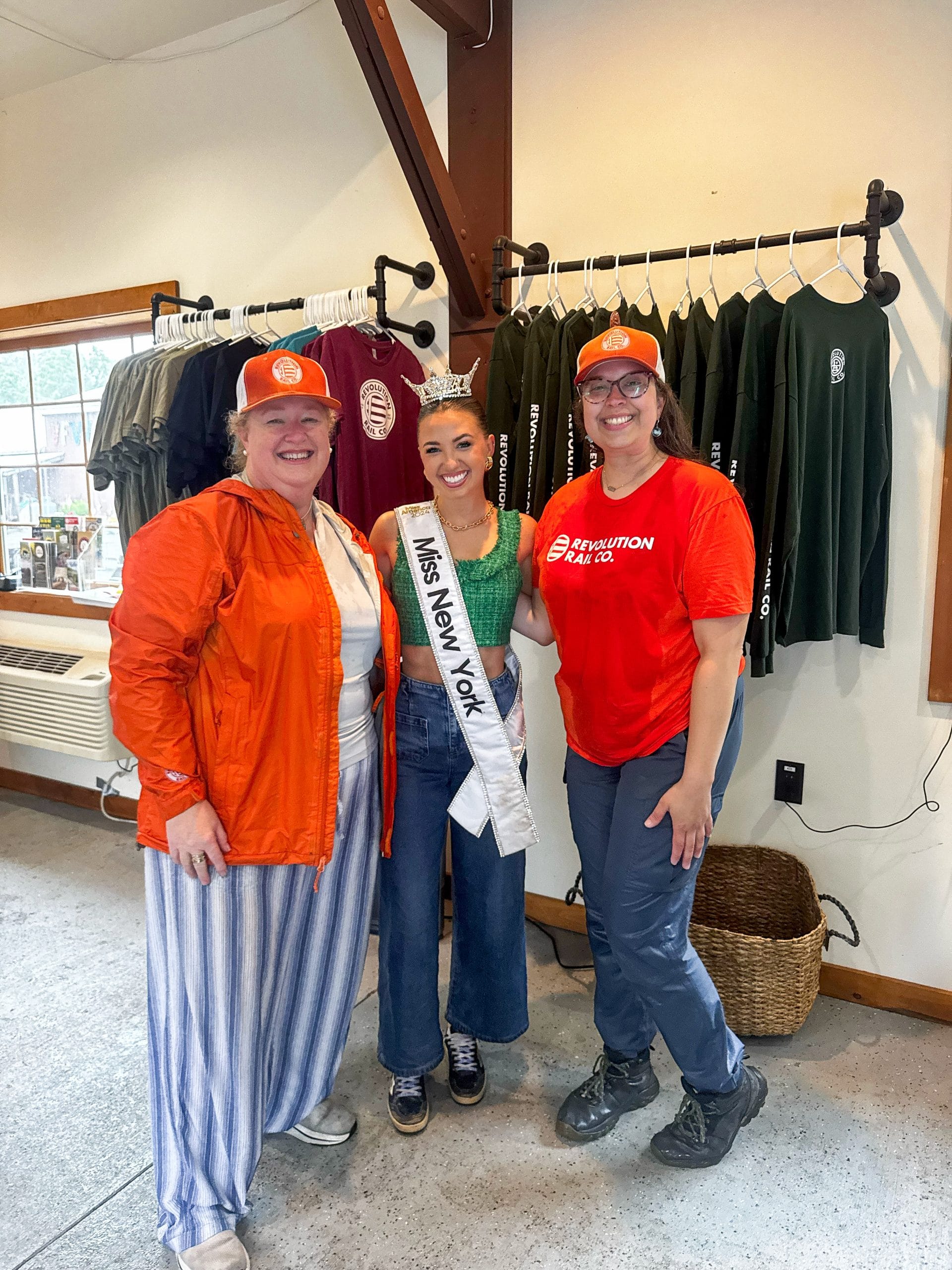 Three women smile indoors at the camera. The woman in the center wears a crown, Miss New York sash, and the other two wear orange hats and casual outfits. Behind them, shirts on display hint at a Lake George event or gathering.