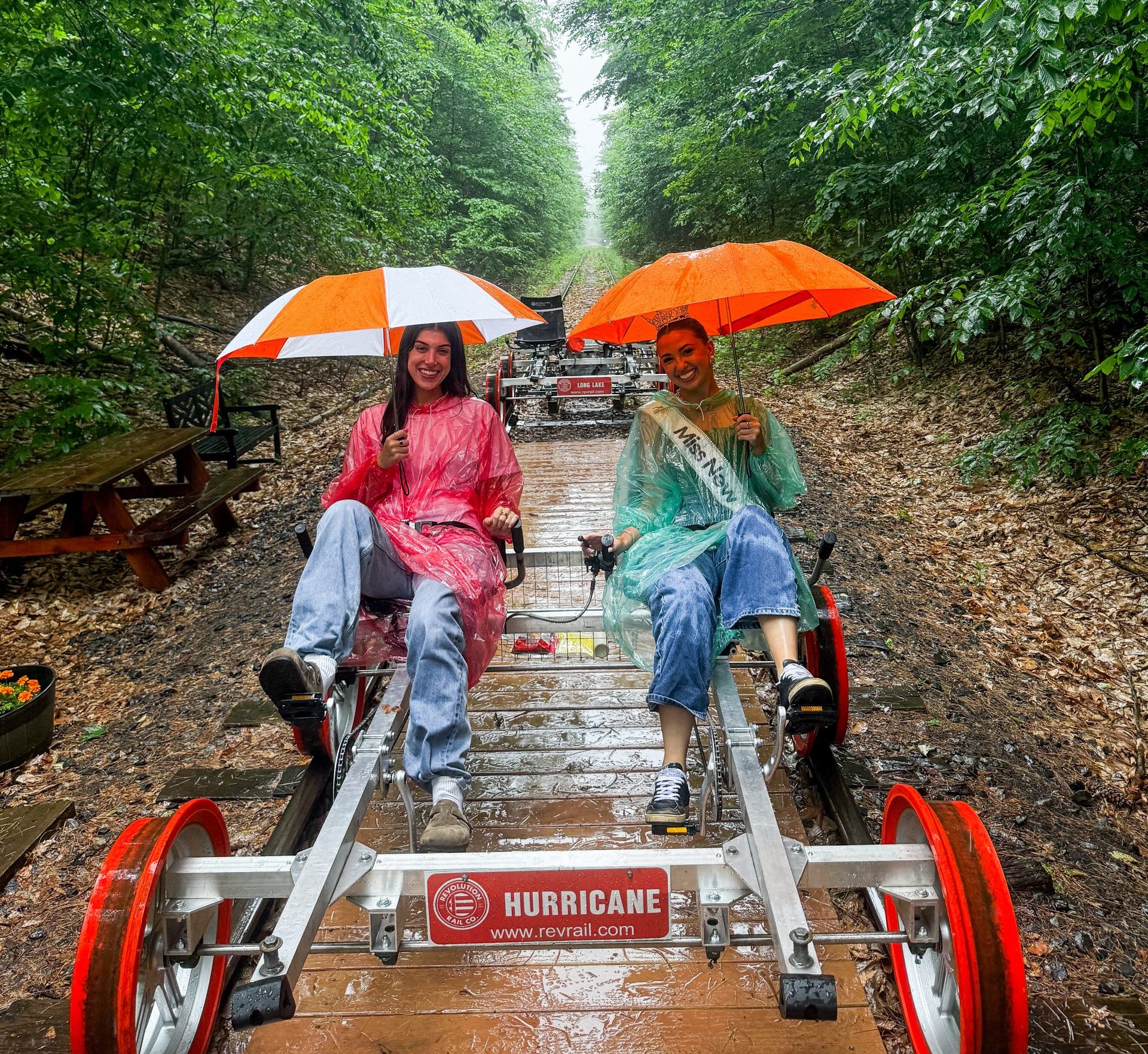 Two people in rain ponchos sit on a pedal-powered rail car holding umbrellas along a wooded, tree-lined trail near Lake George. The wet ground hints at recent rain, and the rail car features a sign reading “HURRICANE” on the front.
