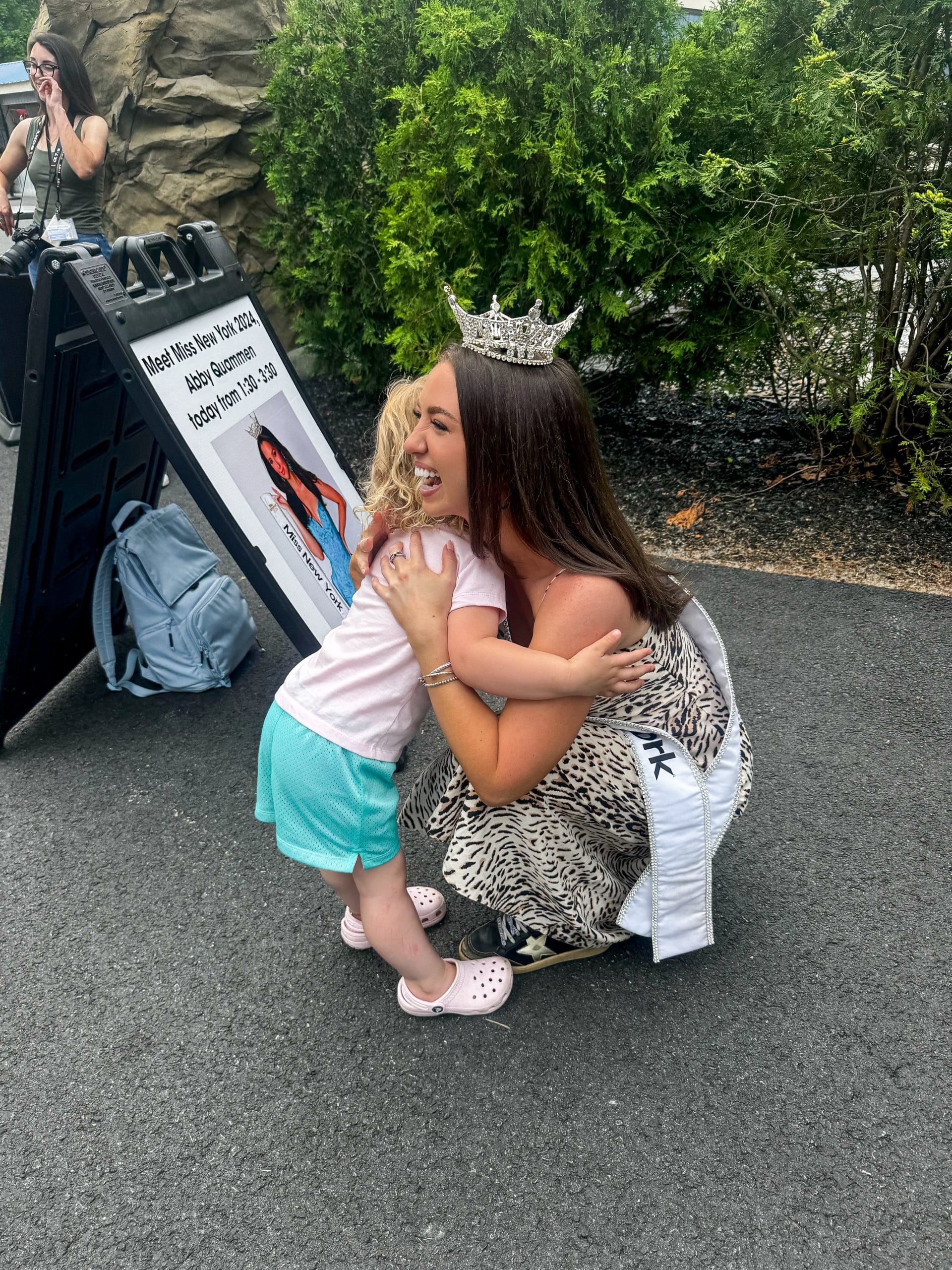 A young woman wearing a crown and sash kneels on a paved path at Lake George, warmly hugging a small blonde-haired child. Greenery and a sign with event details are visible in the background.