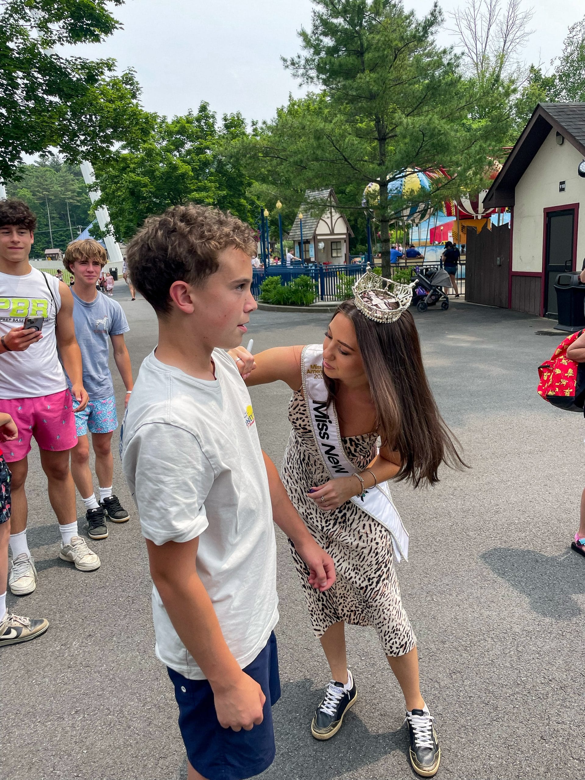 A woman wearing a Miss New York sash and crown smiles and poses with her hand on a boy’s shoulder at an outdoor Lake George amusement park, while other teens stand nearby. Trees and colorful buildings are in the background.