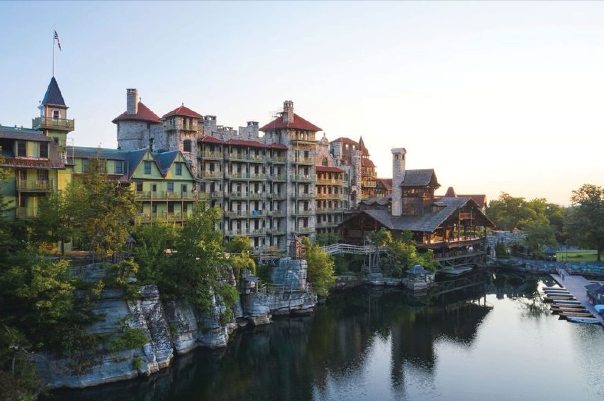 A large historic stone and wood resort hotel with red roofs sits beside a calm lake, surrounded by trees, under a clear sky at sunset—an inviting haven for wellness and relaxation.