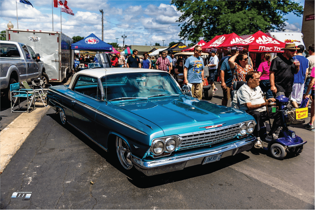 A shiny blue classic Chevrolet car with a white roof is parked at an outdoor Blues Festival, surrounded by people, tents, and vendor booths on a sunny day.