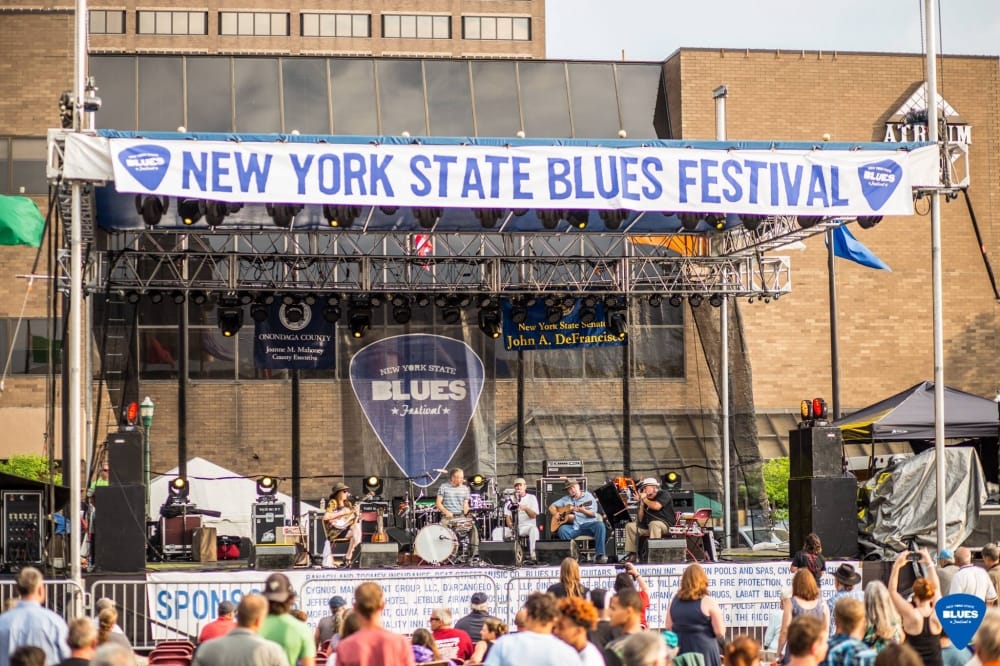A large outdoor stage at the New York State Blues Festival features a band performing live. Similar to the Dutchess County Fair, music lovers gather in front of the stage, while tall buildings rise in the background under a festival banner overhead.
